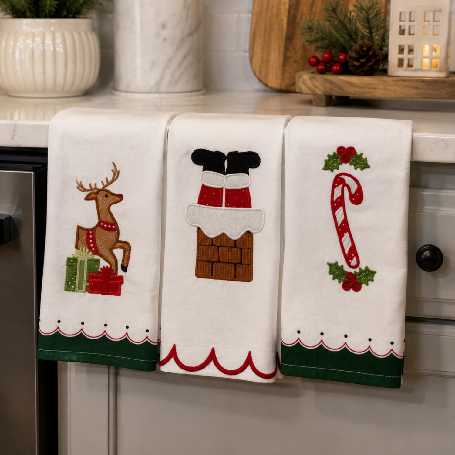 Three white kitchen towels with Christmas-themed embroidery hanging on a countertop. The left towel shows a reindeer with green and red gift boxes, the middle towel shows Santa's boots and chimney, and the right towel shows a candy cane with holly.