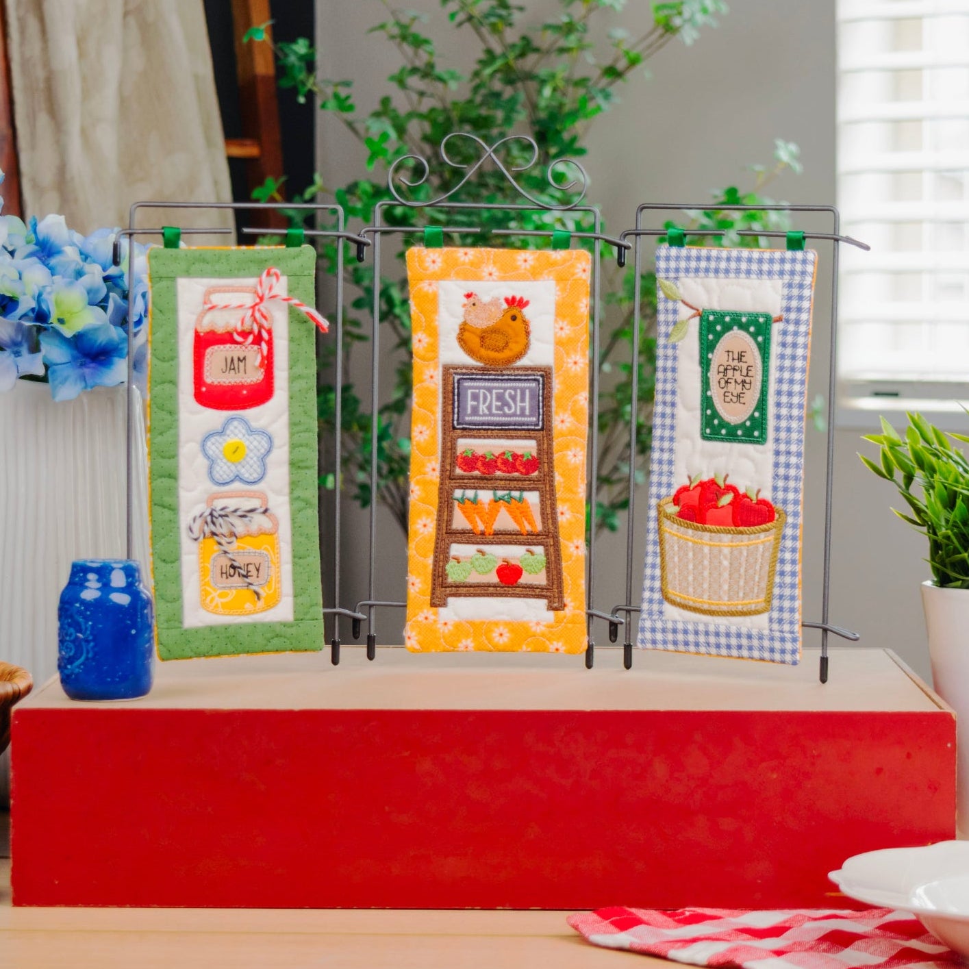 Three decorative kitchen towels displayed on a black wire stand on a red pedestal. The towels feature colorful autumn-themed illustrations including a jar of jam, a pumpkin, and a basket of red apples.