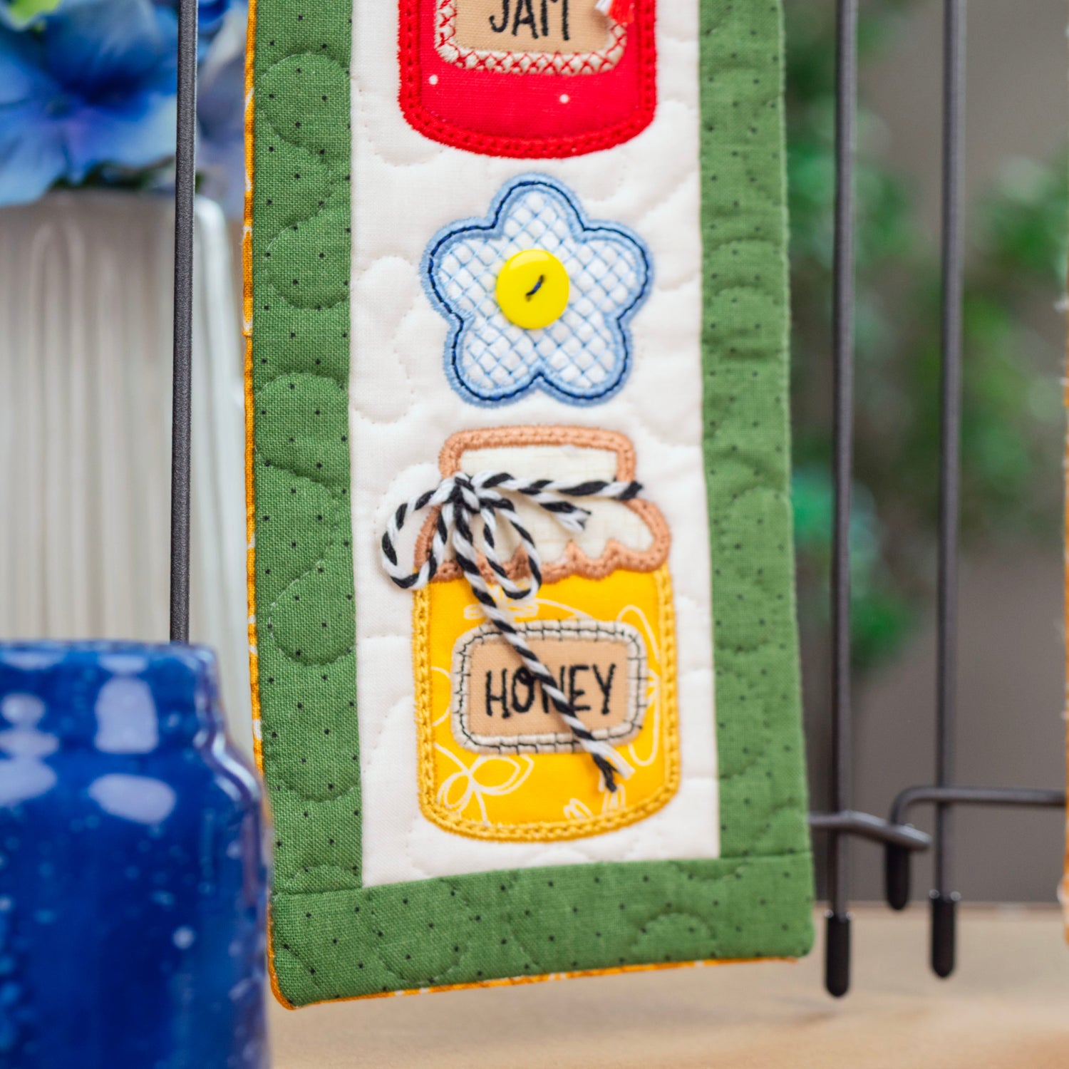 A close-up of a quilted fabric hanging piece with embroidered honey jar, flower, and text details, displayed on a stand alongside other decorative textile pieces.