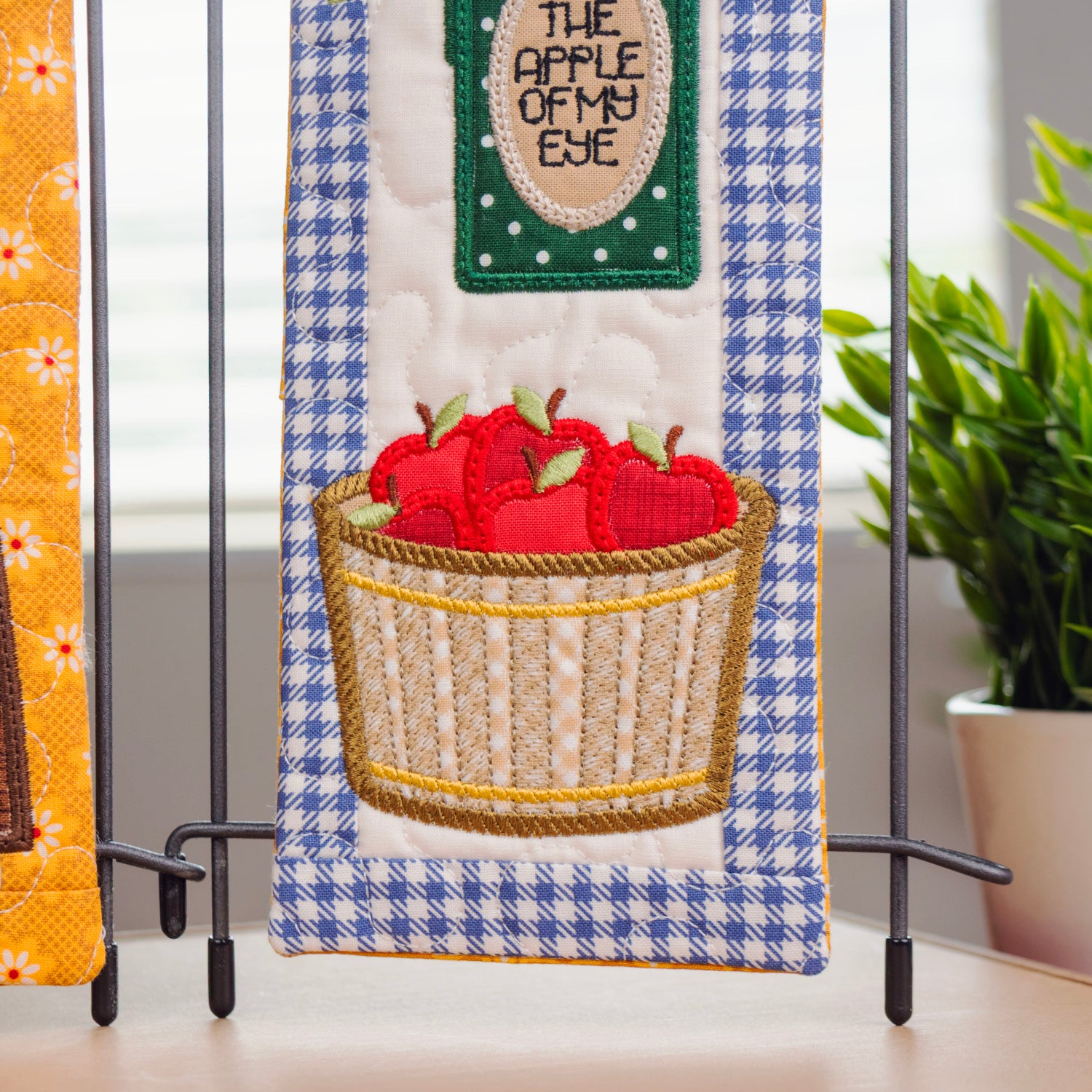 Two decorative quilted fabric wall hangings displayed on a stand in a bright kitchen-like setting. The left hanging shows colorful produce motifs on a yellow gingham background, and the right hanging features red apples in a basket over a blue-and-white gingham panel with a green sign reading 'THE APPLE OF MY EYE.