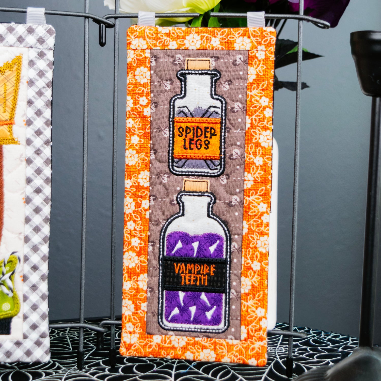 A Halloween-themed decorative sign featuring two glass potion bottles labeled 'Spider Legs' and 'Vampire Teeth' filled with purple liquid, set against a patterned orange and white background. The sign is displayed on a black metal stand on a table with a spiderweb pattern, with dark flowers in the background.