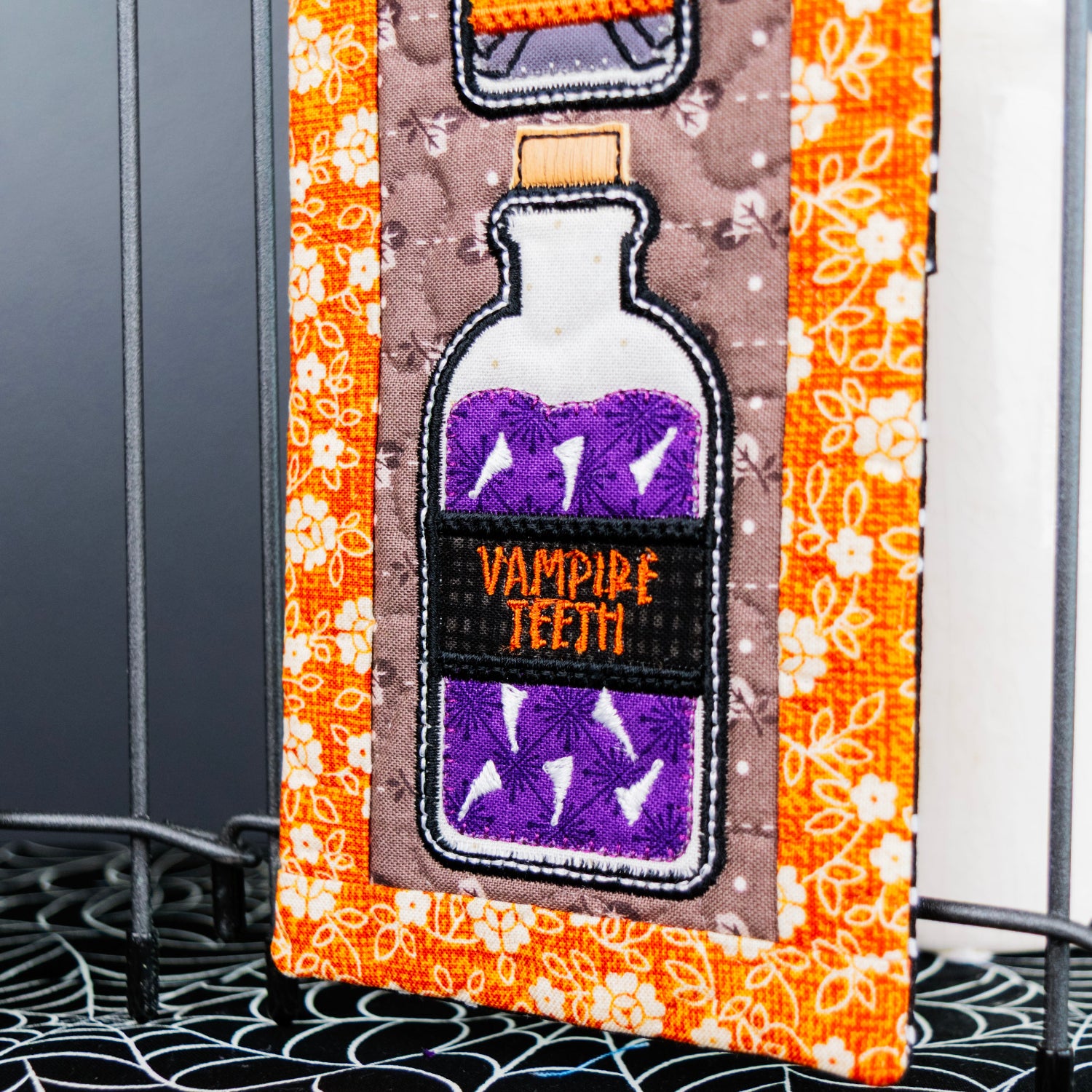 A close-up of a Halloween-themed decorative item featuring an embroidered vampire teeth bottle design in purple and black on an orange patterned background, with part of another similar item visible at the left edge.
