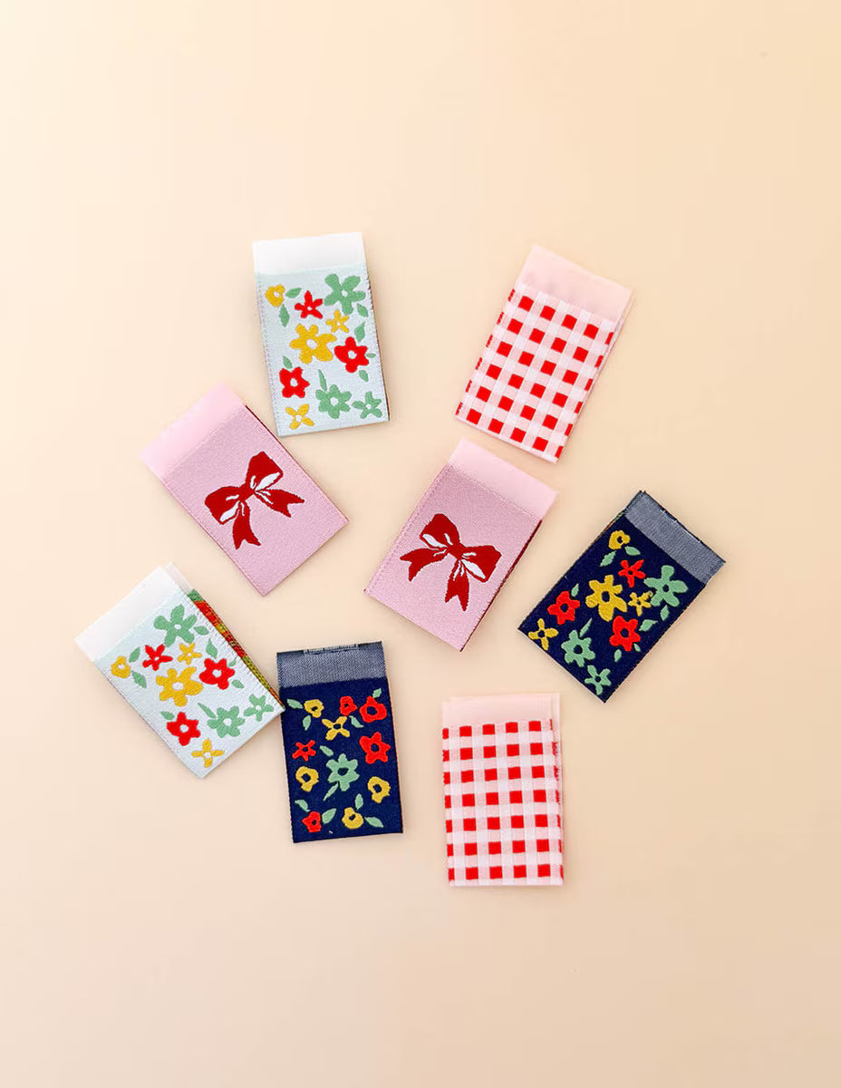 Seven small fabric pouches or card cases arranged on a light beige background. The pouches come in multiple colors and patterns, including pink with a red bow, white with red gingham, navy with multicolor floral prints, and white with multicolor floral prints.