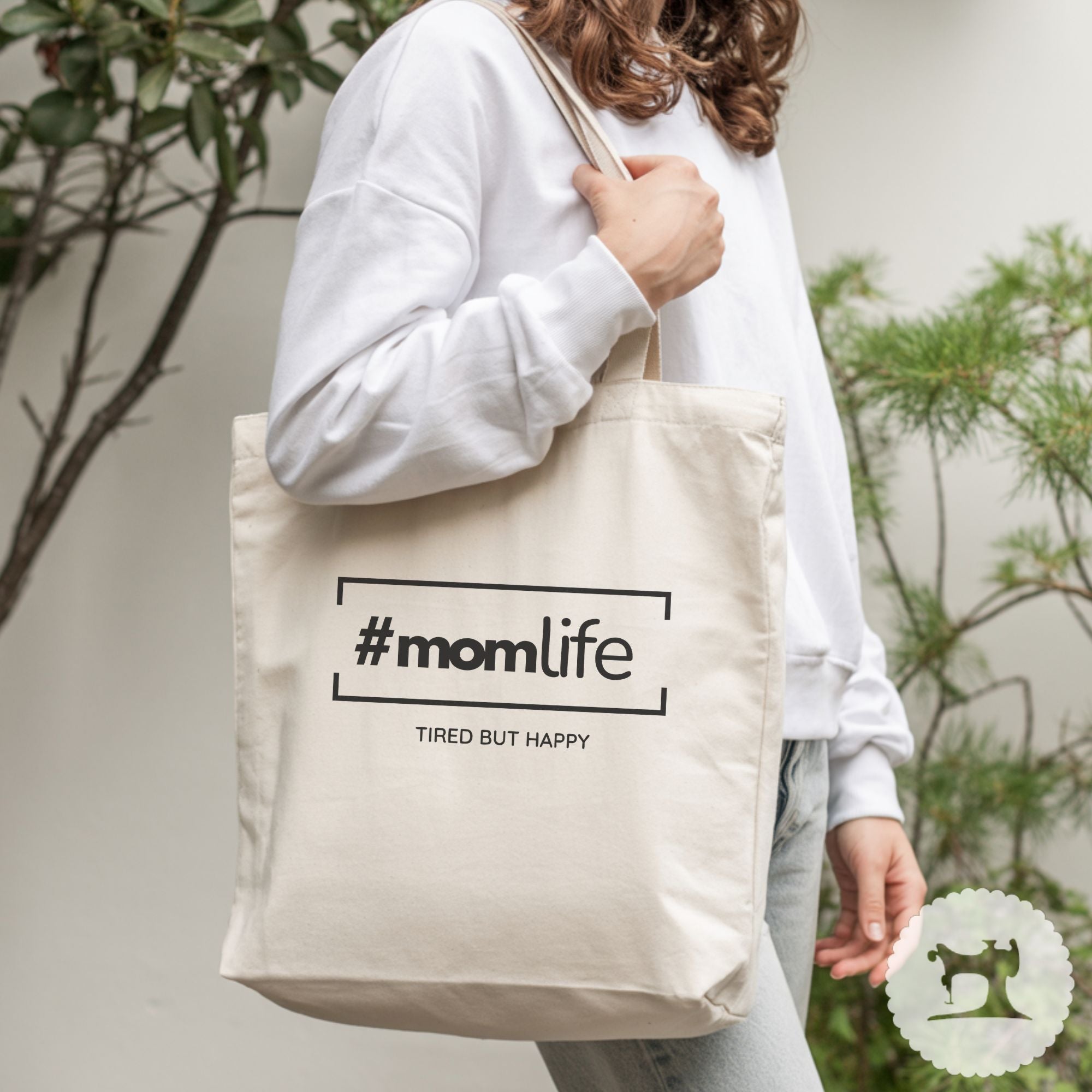 Person holding a tote bag with '#momlife' text, standing outdoors.