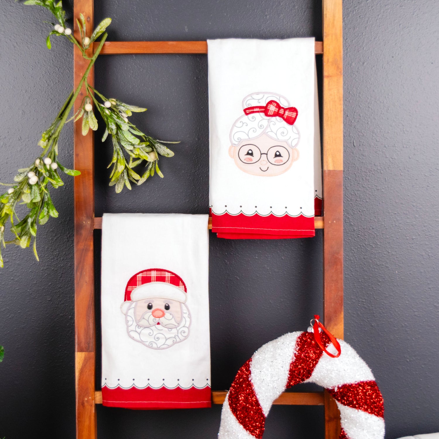 Two white Christmas kitchen towels with embroidered holiday designs hang on a wooden ladder against a dark gray wall. One towel features Santa's face, and the other shows an elderly woman with a red bow and hat.