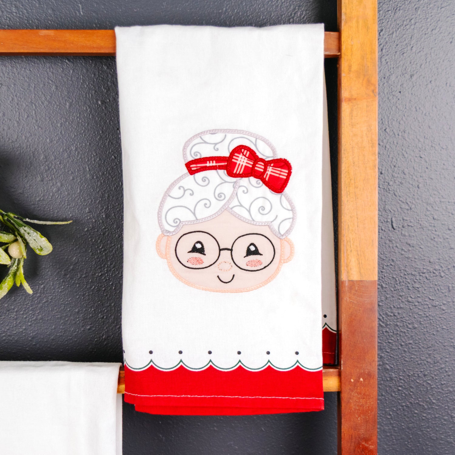 A white kitchen towel featuring an embroidered cartoon granny face with glasses, white hair, and a red bow, hanging on a wooden ladder against a dark gray wall.