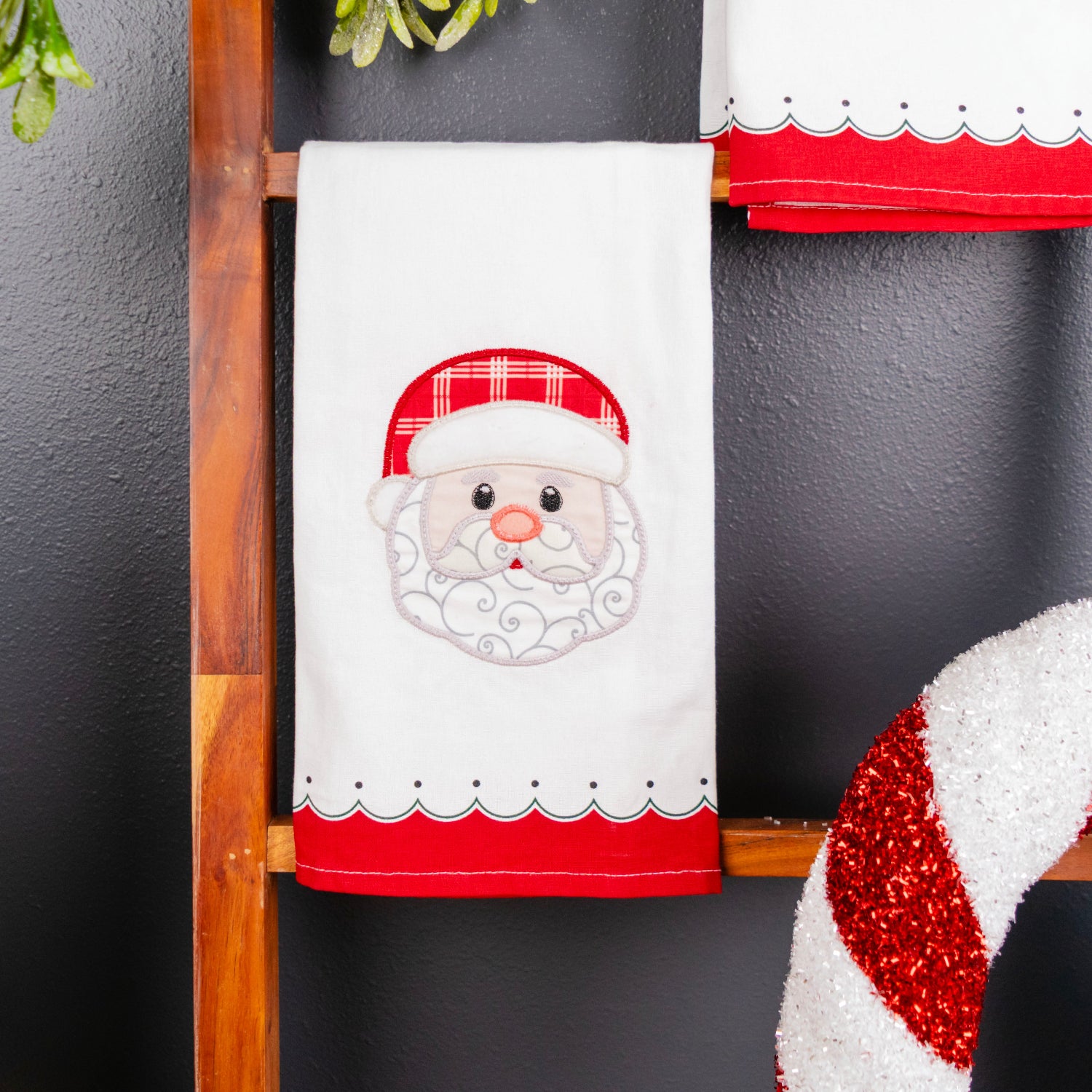A white Christmas kitchen towel with an embroidered Santa face and red hat, hanging on a wooden ladder next to a red and white striped ornament against a dark gray wall.