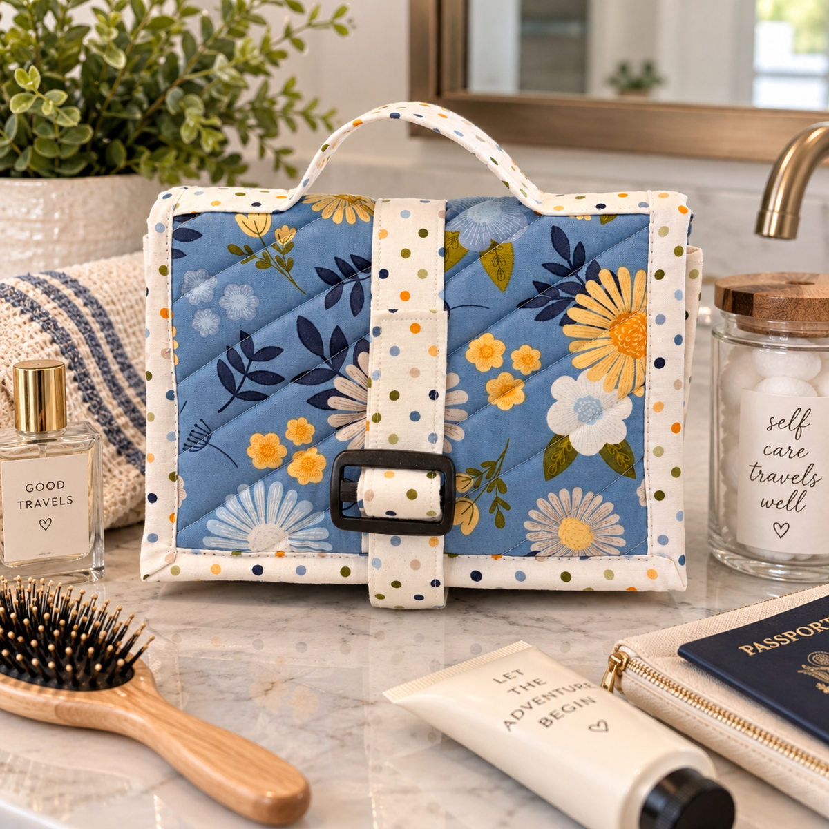A quilted fabric toiletry bag with a blue floral print and white polka-dot trim, styled on a marble bathroom counter with a hairbrush, perfume bottle, lotion tube, and passport nearby.