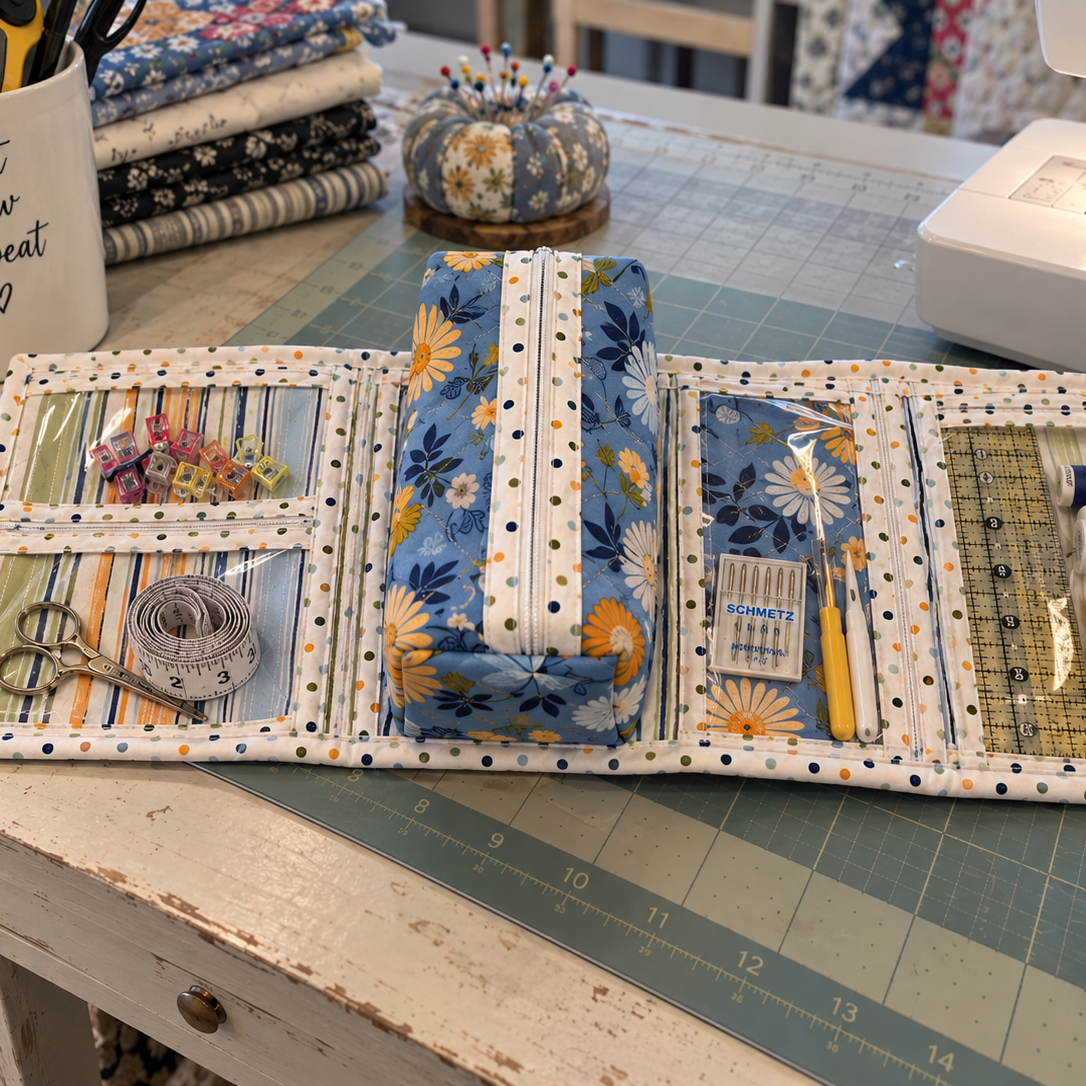 A fabric sewing organizer case laid open on a green cutting mat, holding scissors, thread, pins, ruler, and small accessories. A sewing machine, fabric stack, and sewing supplies are visible around it in a real craft workspace, so this is Lifestyle rather than Product/Packaging.