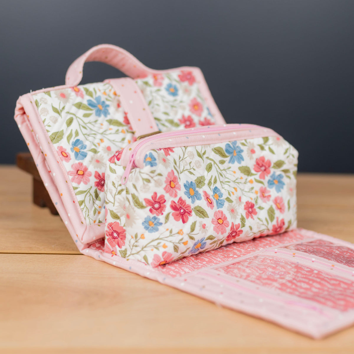 A pink floral zippered pouch with a matching fabric handle, shown partially open on a wooden surface against a dark gray background.