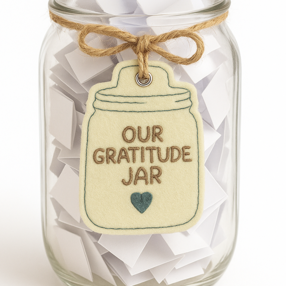 Glass jar labeled 'Our Gratitude Jar' with a string of paper slips inside on a white background