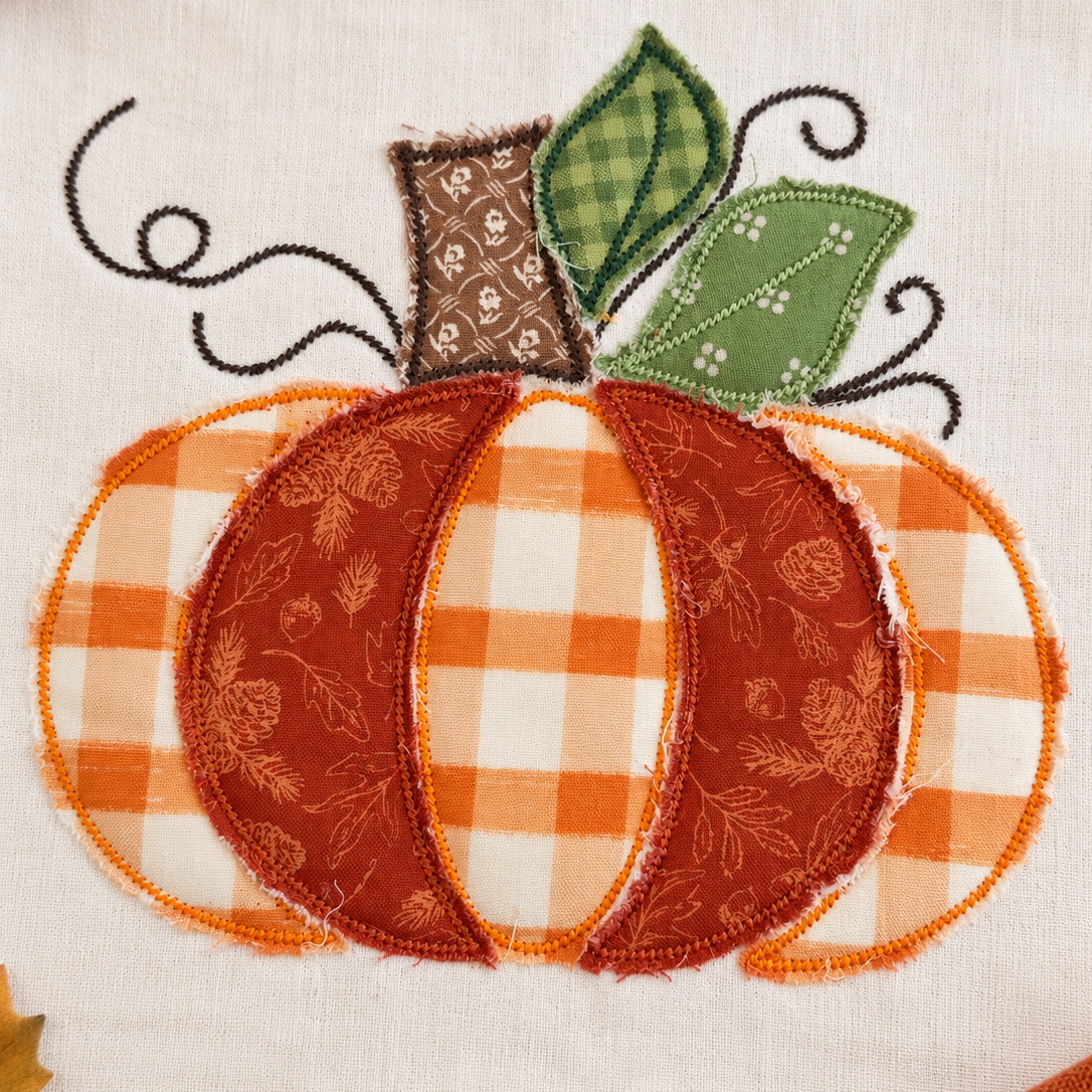 A close-up of a fabric applique pumpkin design on a white textile background, with orange and white gingham sections, a rust-orange patterned center, green leaves, and black vine details. Autumn leaves and small pumpkins are partially visible around the edges.