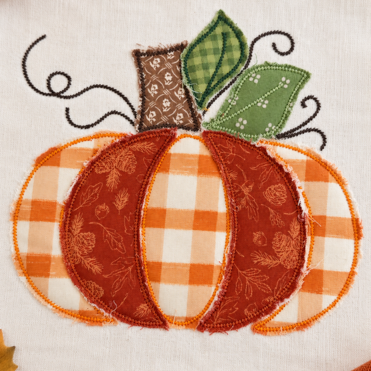 A close-up of a fabric applique pumpkin design on a white textile background, with orange and white gingham sections, a rust-orange patterned center, green leaves, and black vine details. Autumn leaves and small pumpkins are partially visible around the edges.