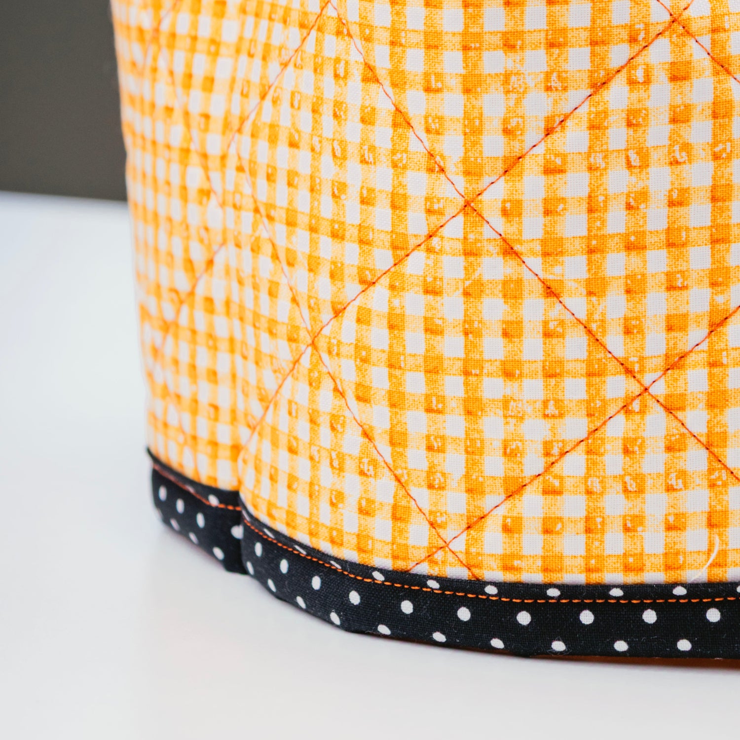 A close-up of a quilted fabric ottoman or pouf with a bright orange and white gingham pattern, finished with a black band of white polka dots along the base.