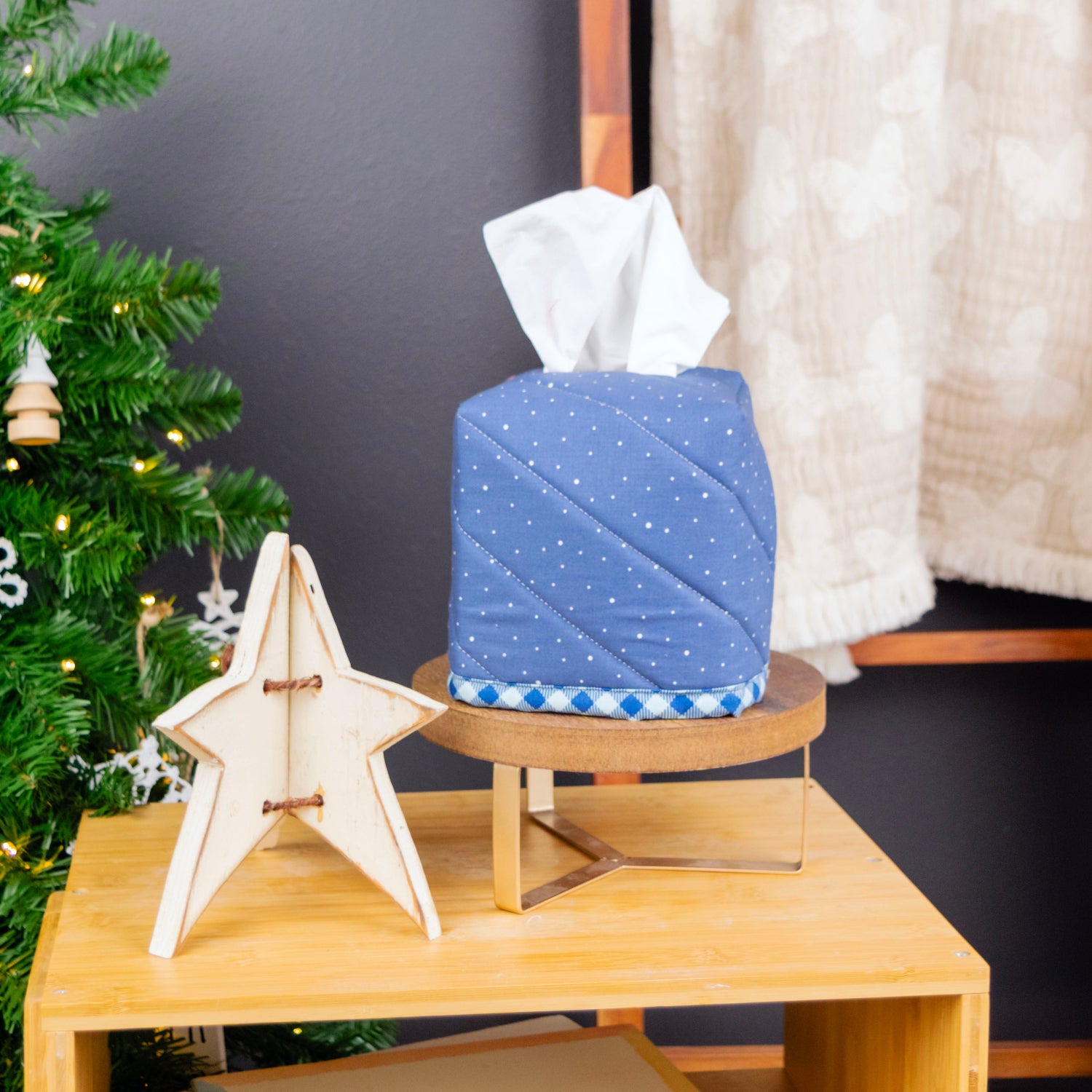 A blue fabric tissue box cover with white polka dots and a checkered trim, holding white tissue paper, displayed on a wooden stand on a light wood shelf in a styled room setting with a Christmas tree and curtain.