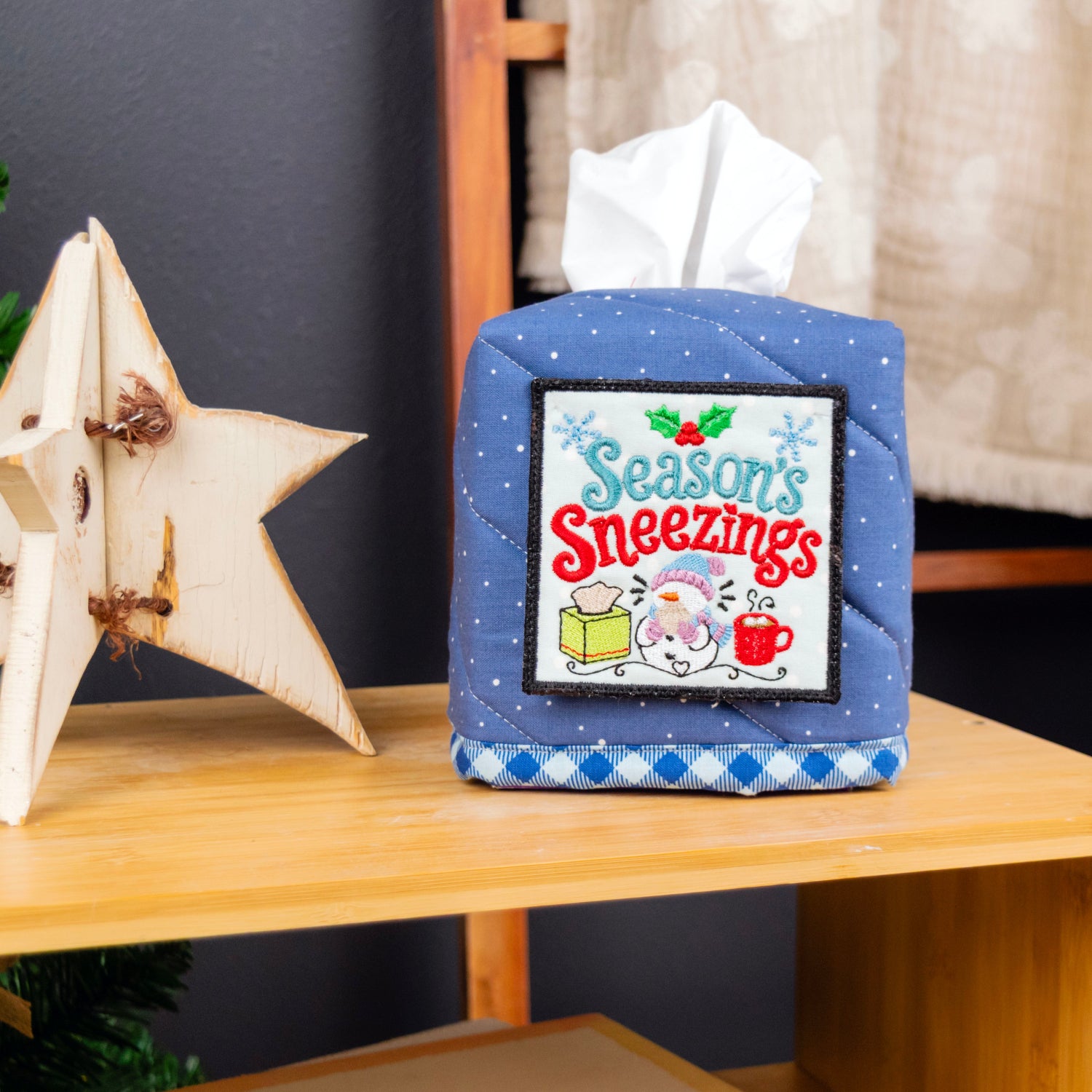 A blue quilted tissue box cover with a festive 'Season's Sneezings' cartoon graphic sits on a wooden table next to a wooden star ornament and a Christmas tree.