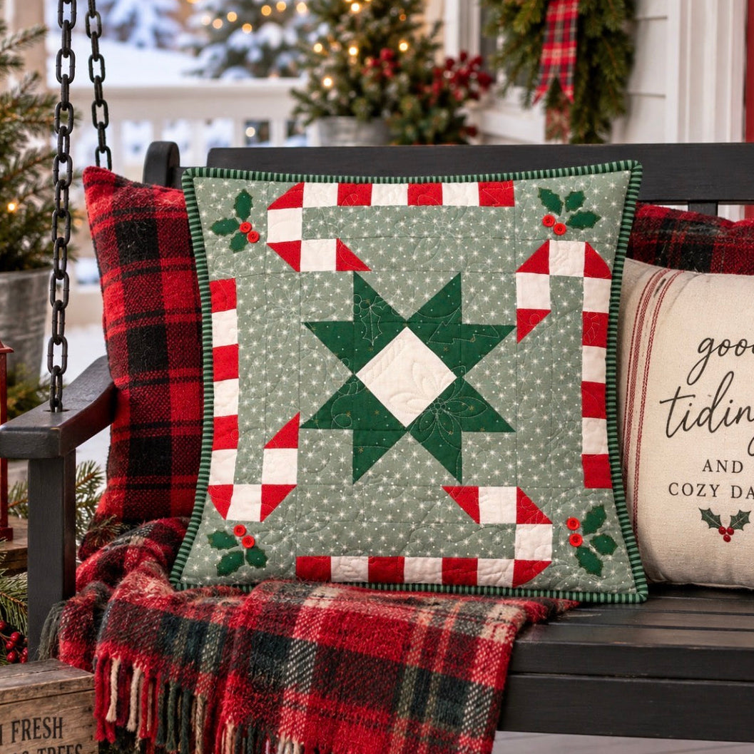 A square quilted Christmas throw pillow with a green star and red-and-white border design, styled on a dark wooden porch bench with a red plaid pillow, a fringed red plaid throw blanket, and a cream pillow that reads 'good tidings and cozy days'.