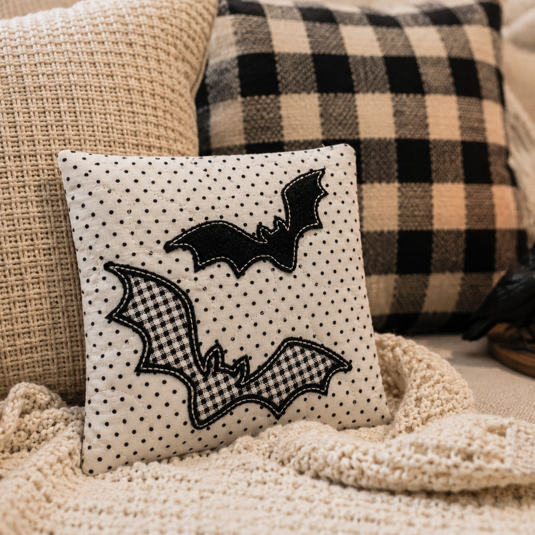 Decorative pillow with bat designs on a couch with plaid pillows and a candle.