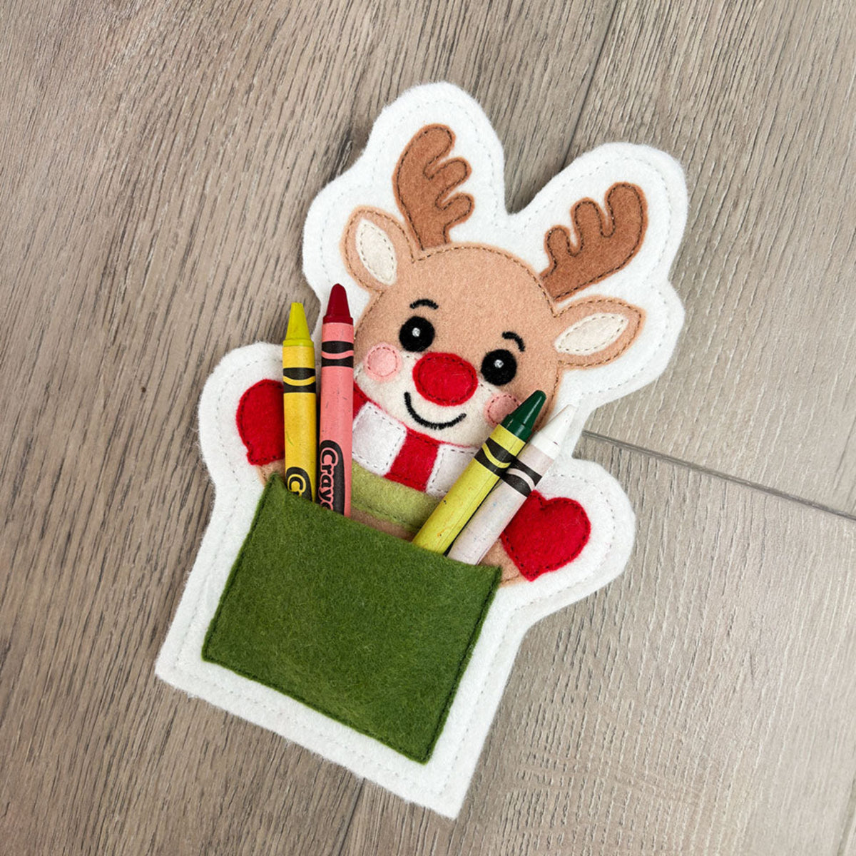 A festive felt reindeer gift holder or pocket on a light wood floor, filled with colorful crayons. The reindeer has brown antlers, a red nose and bow tie, and a green felt pocket holding the crayons.