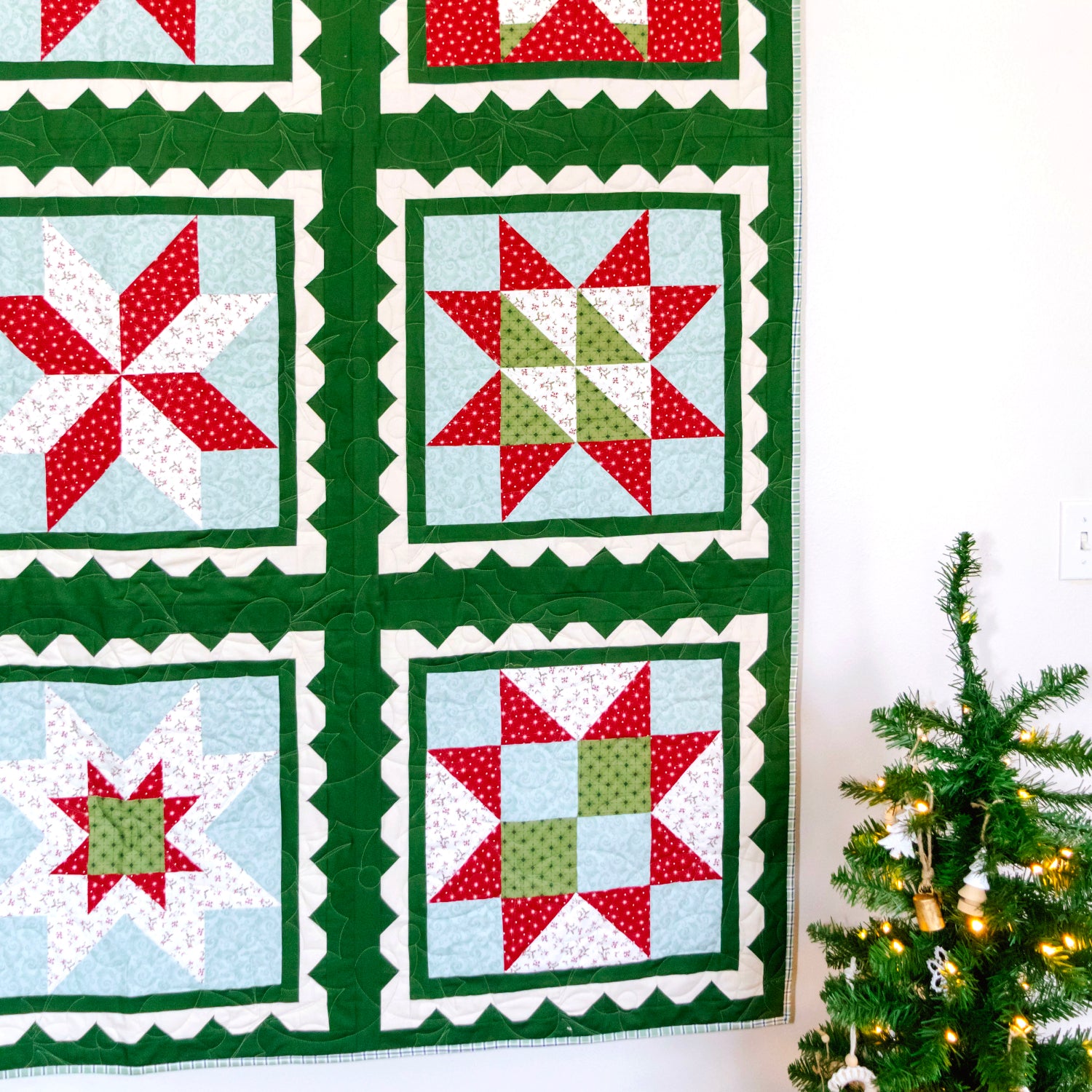 A handmade quilted wall hanging with six Christmas star and snowflake patterns in red, green, and light blue, displayed on a black metal rod against a white wall next to a decorated Christmas tree.
