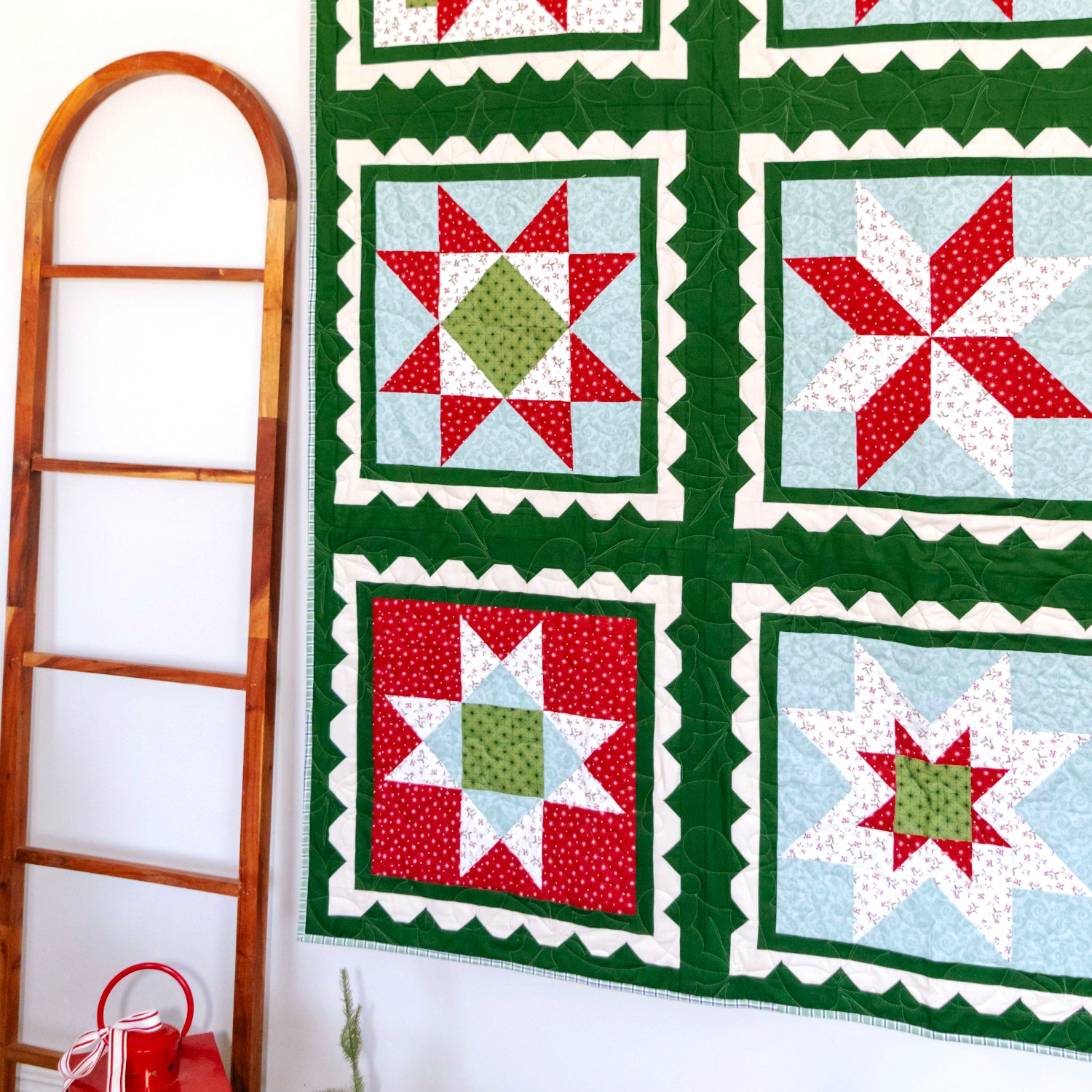 A handmade quilt with six starburst blocks in red, green, white, and light blue, hanging on a black rod against a white wall next to a wooden ladder.