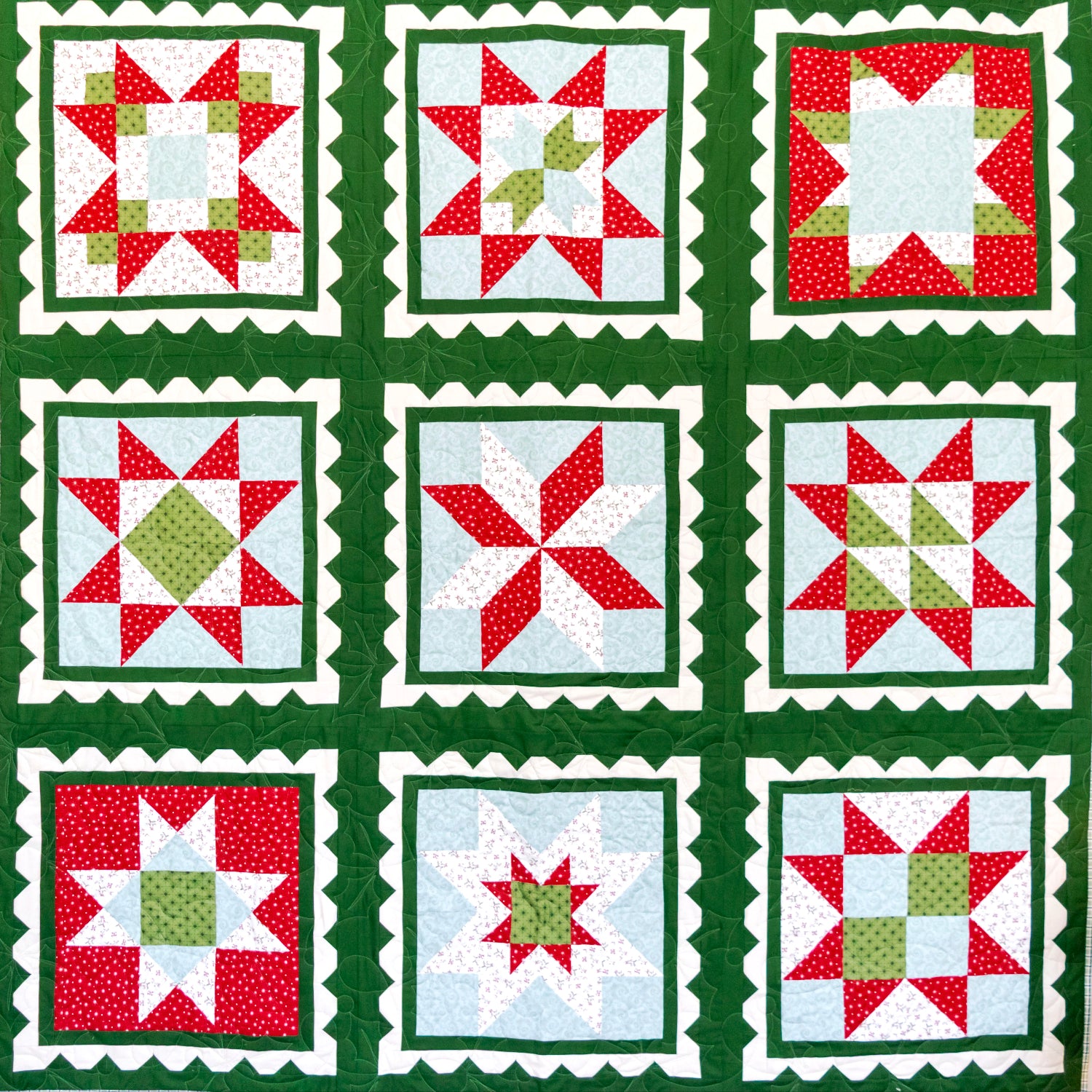 A handmade quilt featuring a 3x3 grid of holiday starburst and geometric blocks in red, green, white, and light blue, hung on a white wall next to a wooden ladder and a small Christmas tree.