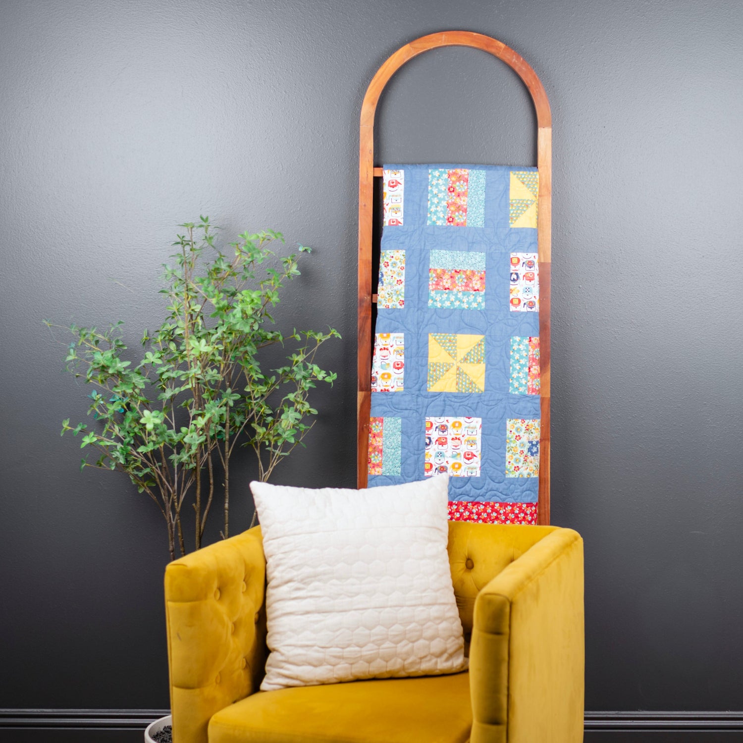 A colorful patchwork quilt hanging from an arched wooden rack, styled in a room setting with a mustard yellow armchair, a white pillow, and a potted plant against a dark gray wall.