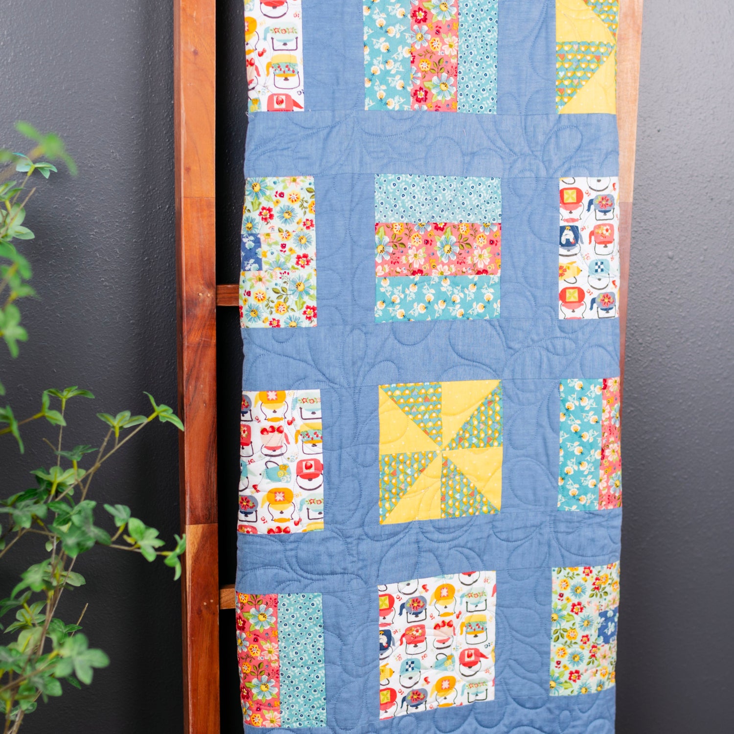 A handmade quilted fabric panel with a blue background and colorful patchwork blocks, hanging on a wooden hanger against a dark gray wall with green foliage on the left.