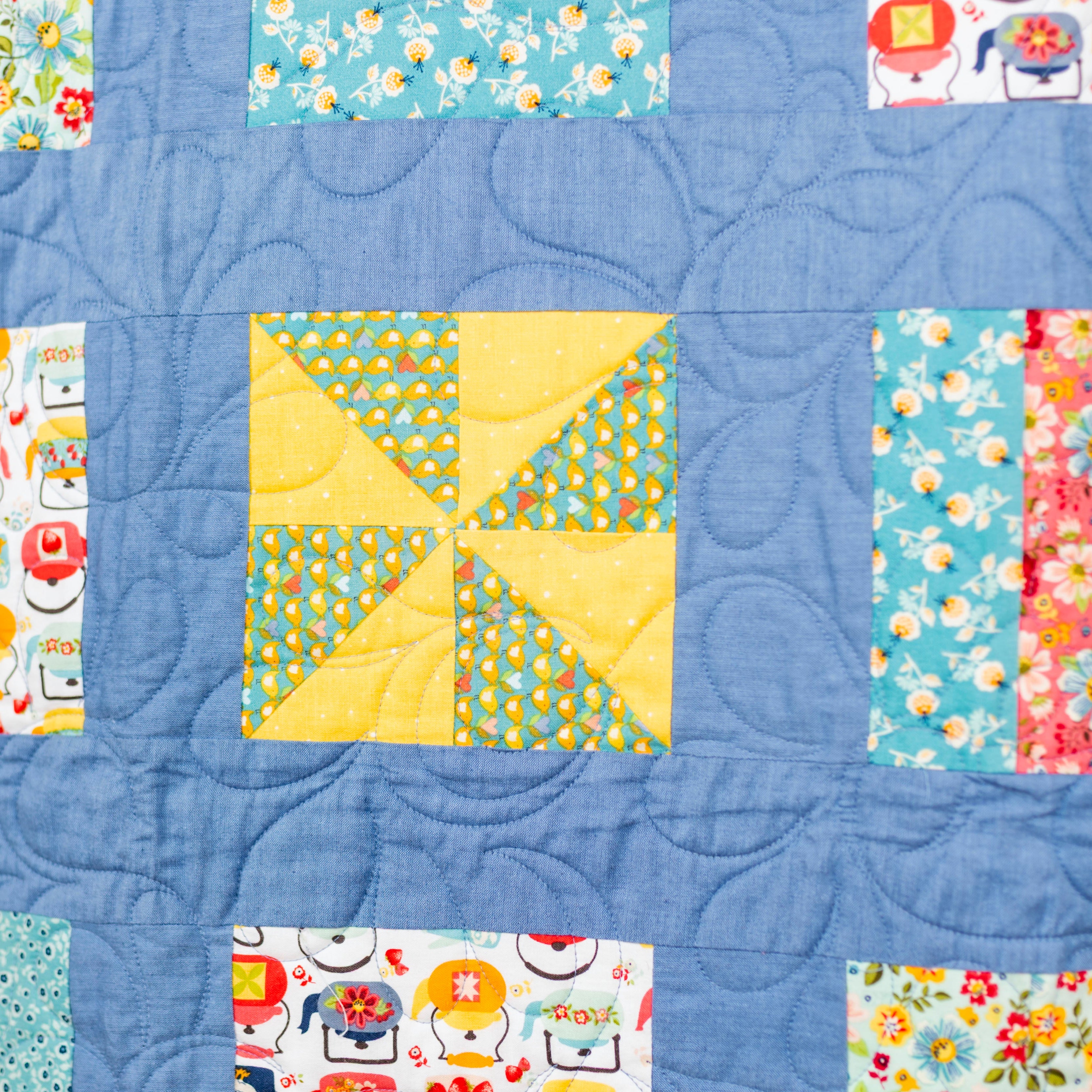 A close-up of a handmade quilted textile featuring a central yellow and blue geometric block surrounded by various colorful floral and patterned fabric borders on a blue background.