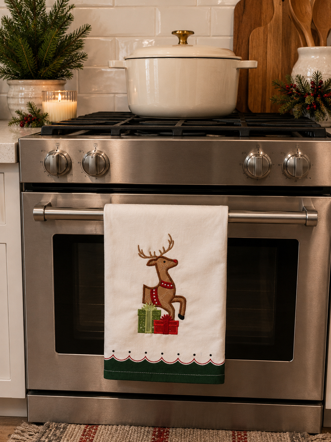 A white kitchen towel with an embroidered brown reindeer and green/red gift box design hanging on the handle of a stainless steel gas range. The oven door is closed, with a white Dutch oven on the stovetop and holiday decor in a kitchen setting.