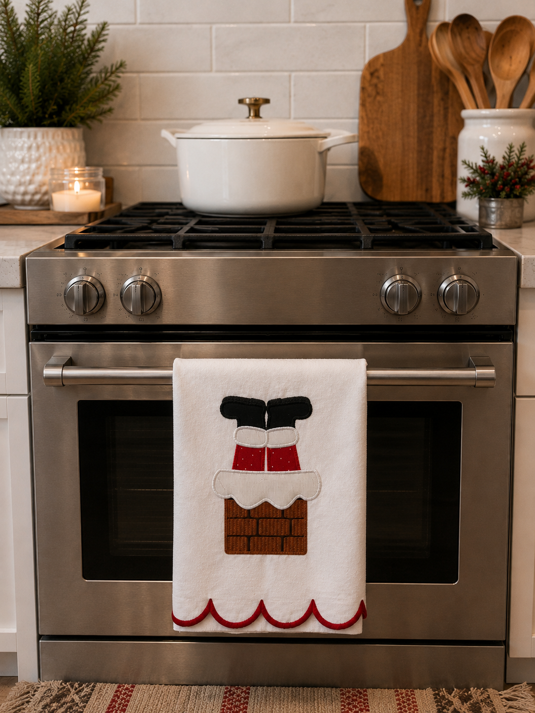 A white kitchen towel hanging from the handle of a stainless steel freestanding range, featuring an applique design of Santa's boots in a chimney stack. The background shows a white subway tile backsplash, a white pot on the stovetop, and wooden kitchen utensils.