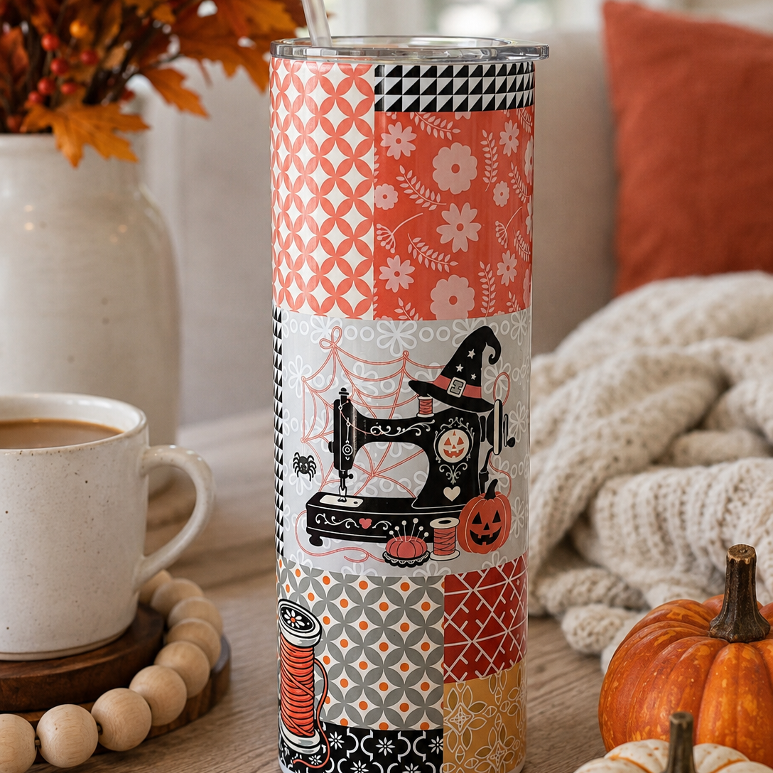 A tall insulated tumbler with a clear lid and metal straw, decorated with a patchwork of orange, black, white, and beige autumn-themed patterns including a sewing machine, pumpkins, and floral motifs. The tumbler is styled on a wooden table with a coffee cup, a vase of fall leaves, a knitted blanket, and small pumpkins.