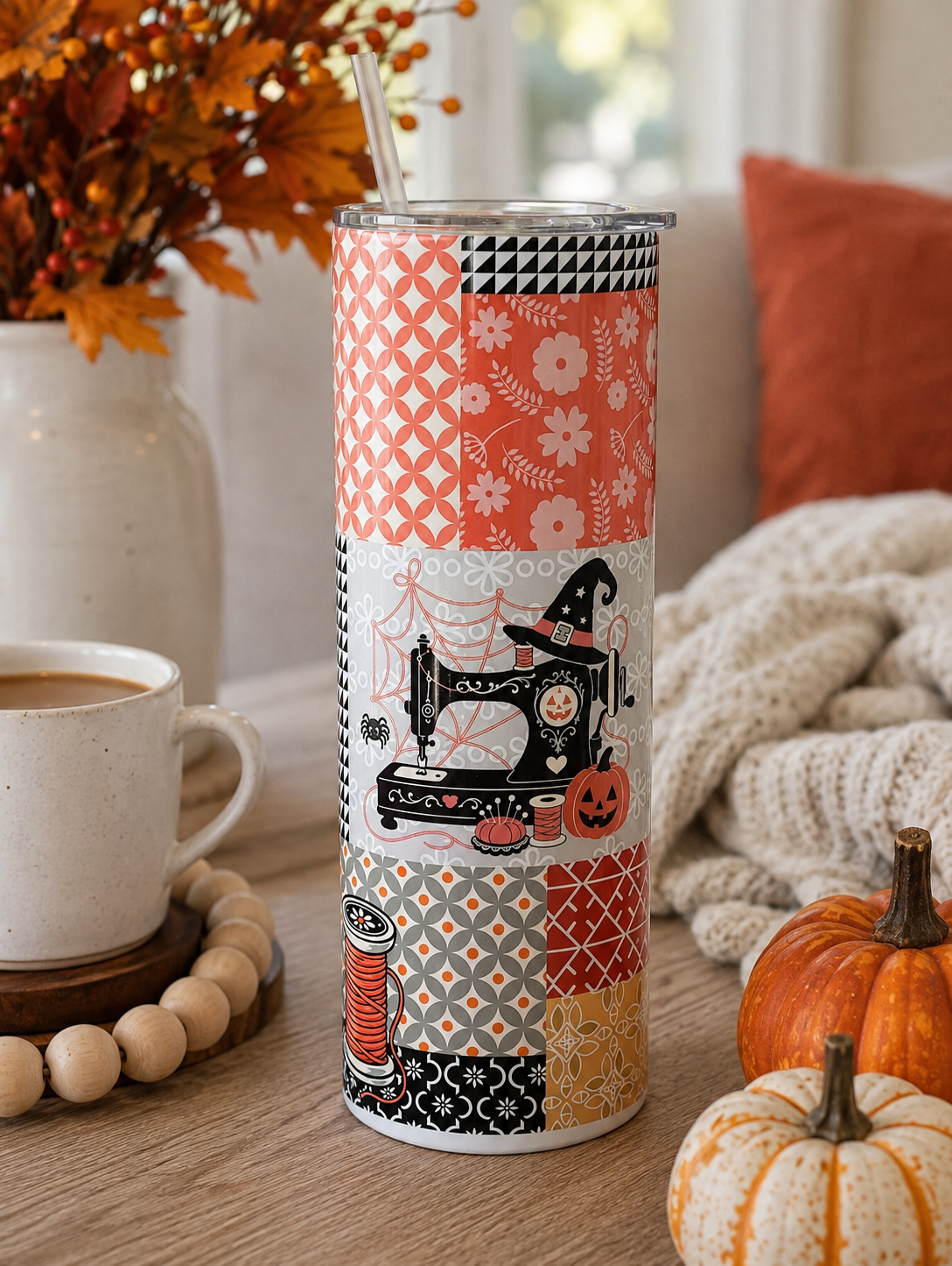 A tall insulated tumbler with a clear lid and metal straw, decorated with a patchwork of orange, black, white, and beige autumn-themed patterns including a sewing machine, pumpkins, and floral motifs. The tumbler is styled on a wooden table with a coffee cup, a vase of fall leaves, a knitted blanket, and small pumpkins.