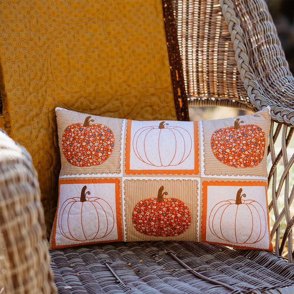 A rectangular decorative throw pillow with a 3x2 grid pumpkin pattern, styled on a wicker patio chair next to a yellow cushion.