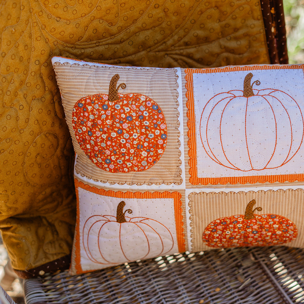 A rectangular decorative throw pillow with a patchwork autumn pumpkin design, resting on a wicker chair next to a yellow quilted cushion.