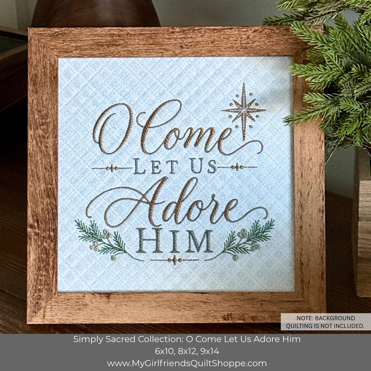 A framed decorative sign with the phrase “O Come Let Us Adore Him” in brown script and block lettering on a light blue quilted background. The sign is housed in a rustic brown wooden frame, styled with evergreen branches on a dark wooden surface.