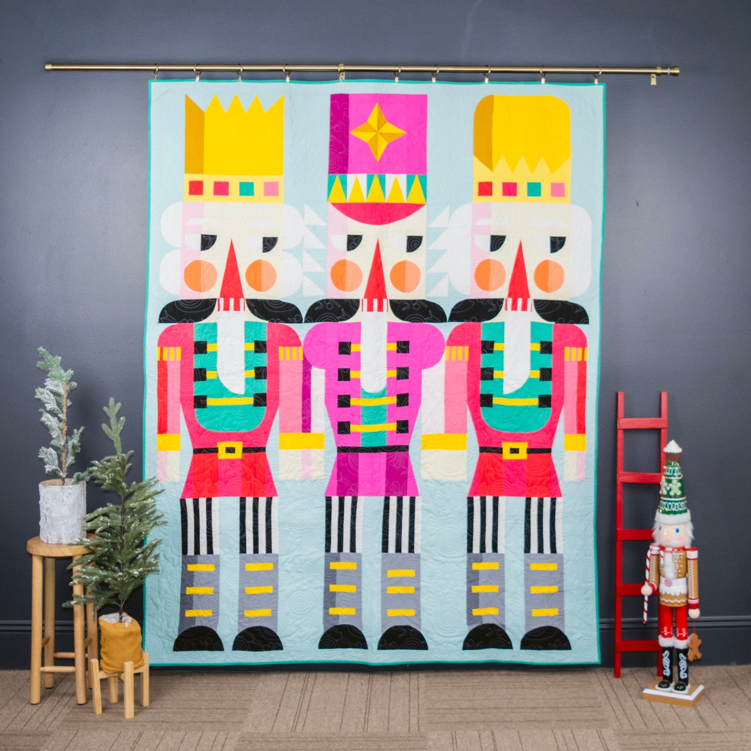 A large textile wall hanging featuring three colorful nutcracker figures on a light blue background, displayed in a styled room with a dark gray wall, wood floor, small plants on a stand, and a red ladder.