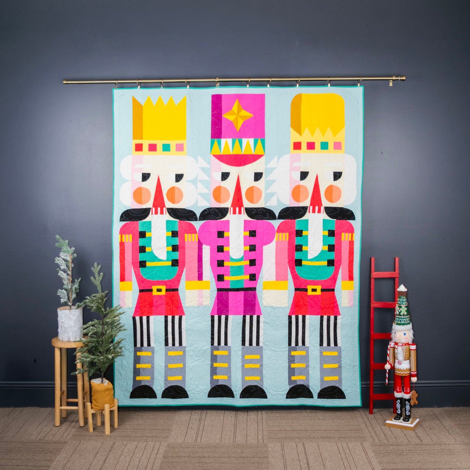 A colorful quilt featuring three stylized nutcracker figures with bright pink, red, teal, black, gray, and yellow blocks on a light blue background, displayed indoors.