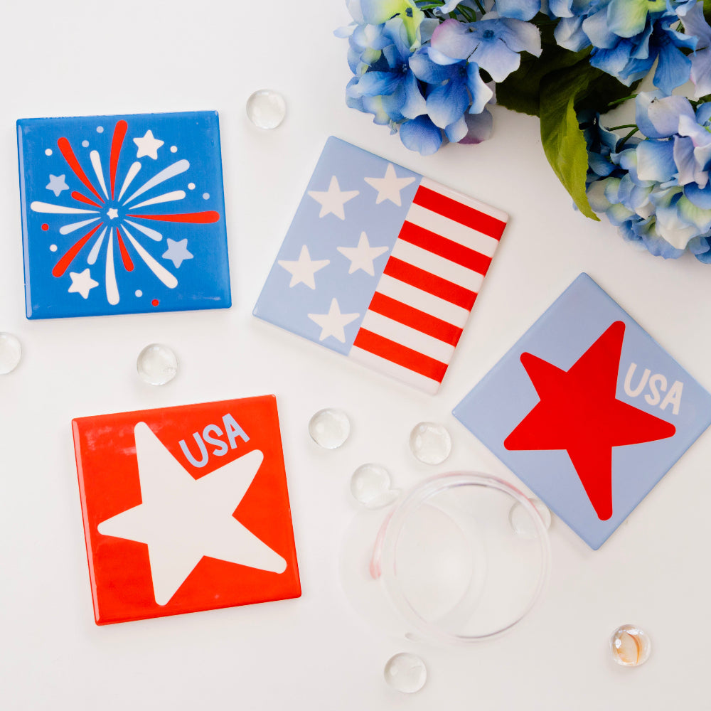 Four square coasters with patriotic designs on a white surface with blue flowers.