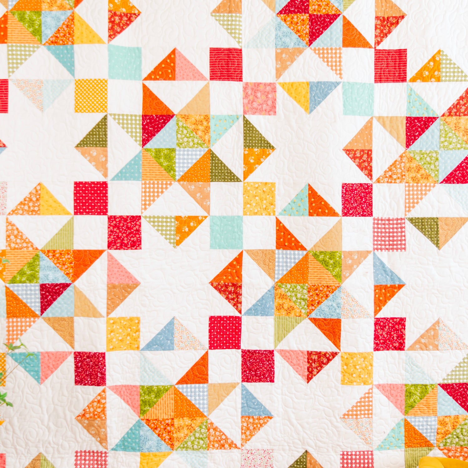 A colorful patchwork quilt hanging on a dark gray wall, featuring a geometric starburst pattern with triangles and squares in orange, red, yellow, green, and light blue on a white background.
