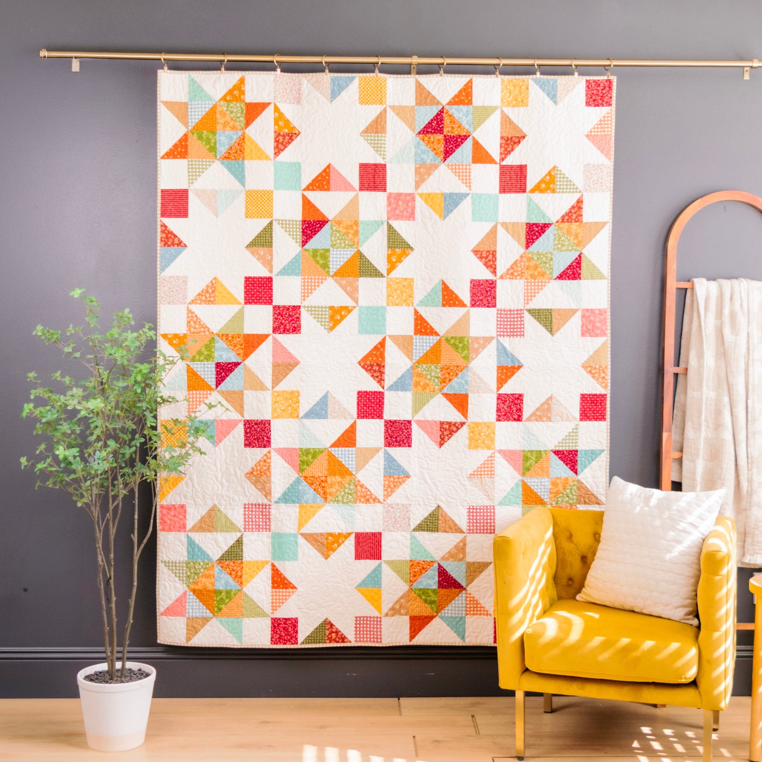 A colorful geometric quilt hanging on a wall in a styled room, with a yellow armchair, potted plant, and wooden ladder shelf nearby.