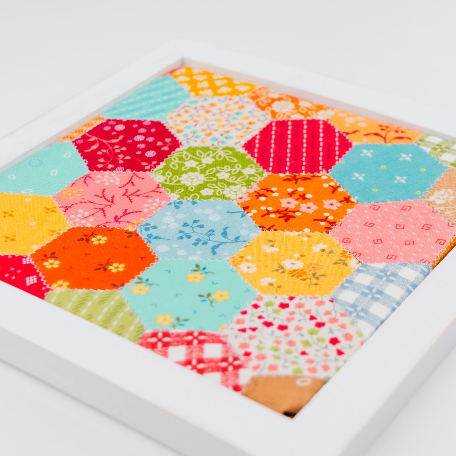 A square white framed art piece featuring a colorful patchwork quilt design made of hexagonal fabric blocks in various patterns and colors, photographed on a clean white background.