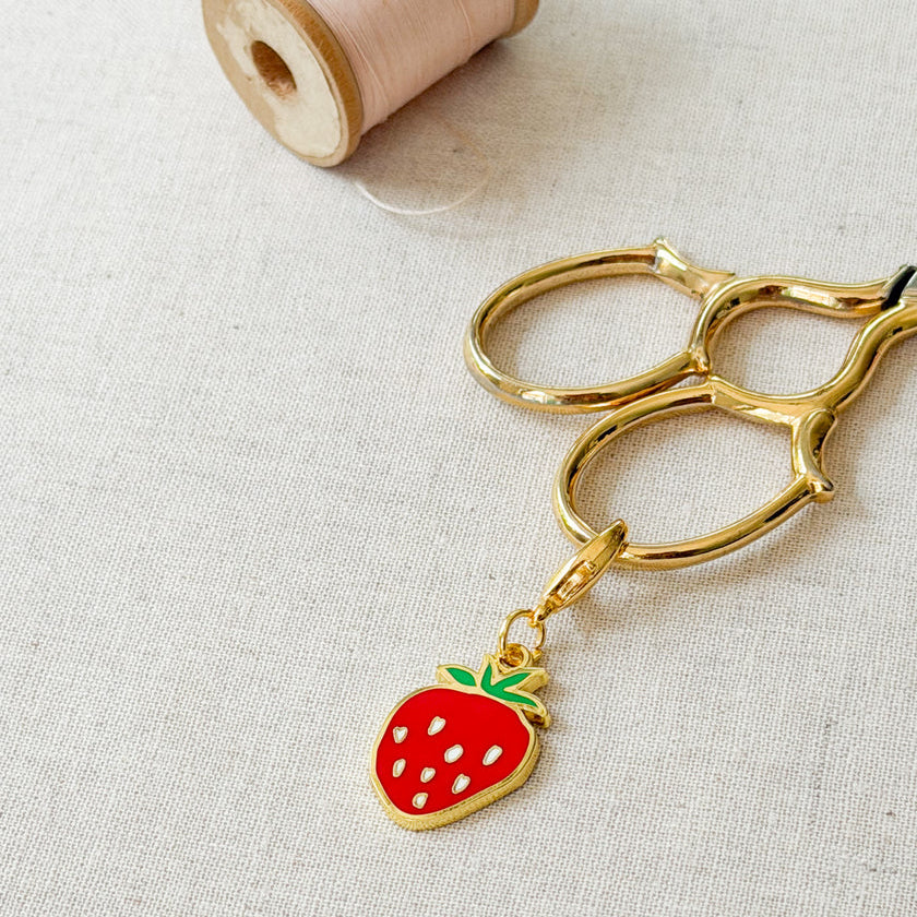 A gold metal key ring with a dangling strawberry charm, placed on a light beige linen surface next to a spool of pink thread.