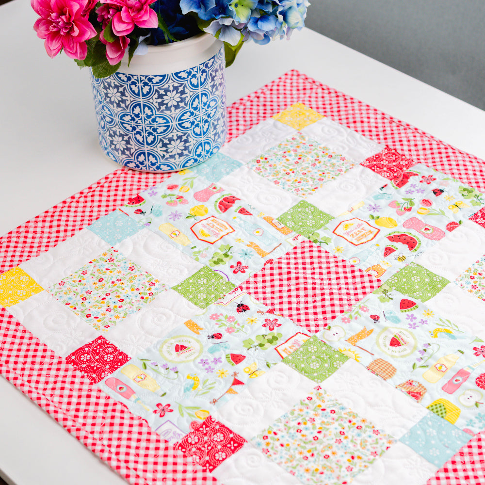 Quilted table runner with colorful patterns on a white table, accompanied by a vase of flowers.