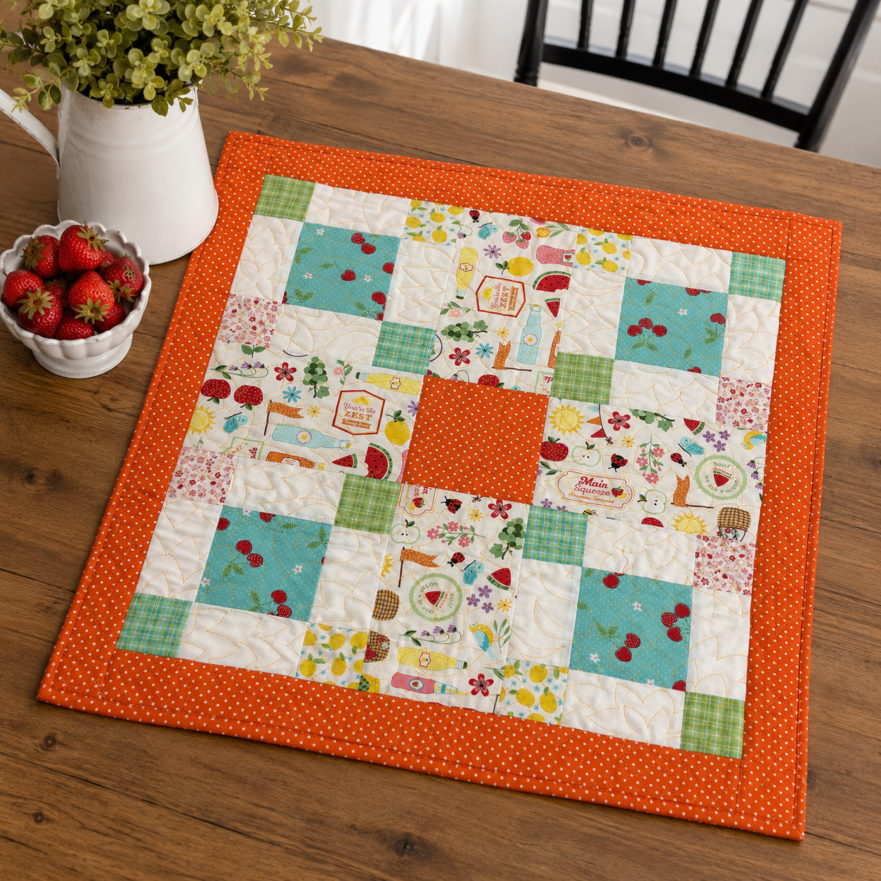 A square quilted patchwork placemat with an orange border and a central orange square, displayed on a wooden table. The patchwork design features various colorful retro-themed patterns including cherries, lemonade stands, and floral motifs.