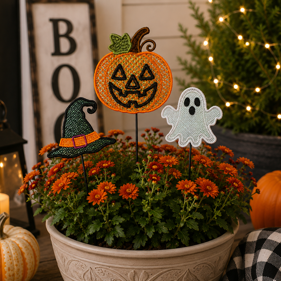 Halloween-themed floral garden stake decorations featuring a jack-o'-lantern, a witch hat, and a ghost, displayed in a pot of orange mums with decorative pumpkins and a lit Christmas tree in the background.