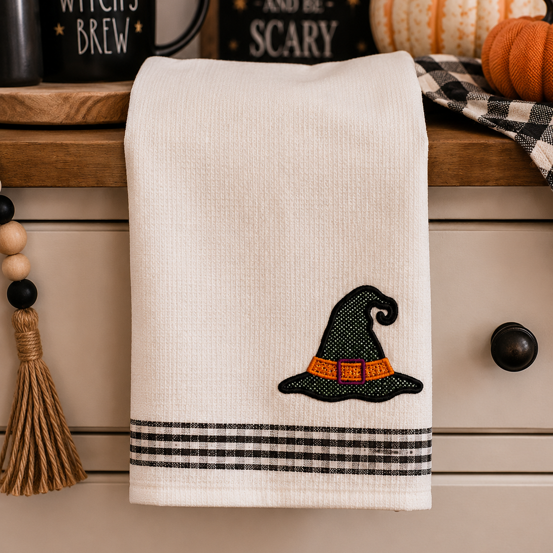 An off-white cotton kitchen towel with a black witch hat and orange band embroidery, plus a black-and-white plaid stripe border at the bottom, hanging on a dresser in a fall-themed setting.