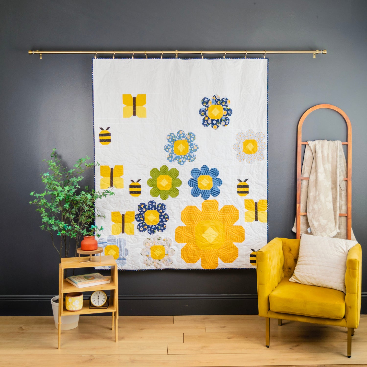 A white fabric wall hanging or curtain featuring a colorful floral and bee pattern, displayed on a gold rod in a styled room with a yellow armchair, side table, and dark gray wall.