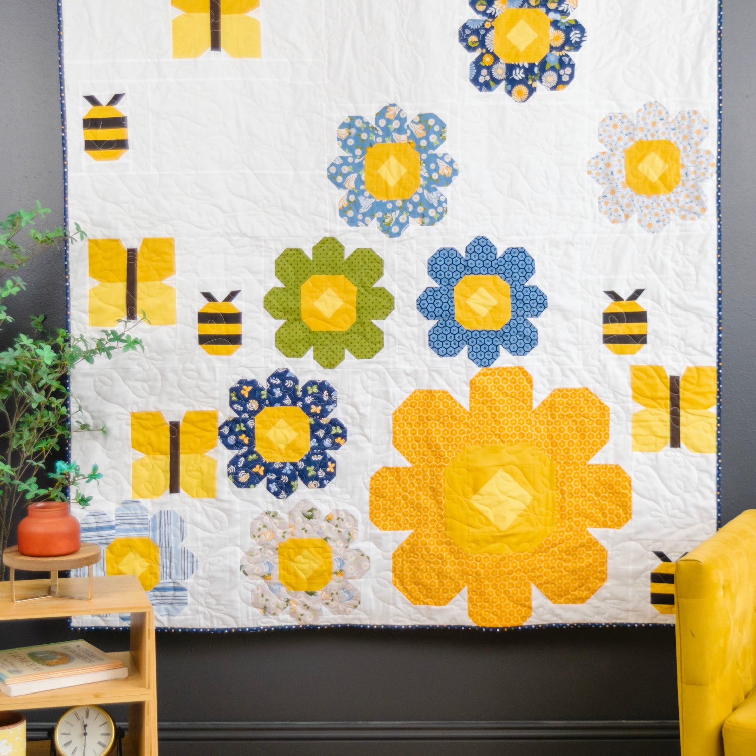 A colorful quilted wall hanging or small quilt displayed in a styled room corner, featuring large yellow flowers, blue and green flower motifs, and bumblebee appliqués on a white background. The room includes a dark gray wall, a yellow armchair, a wooden side table with books and a plant, and a peach-framed chair.