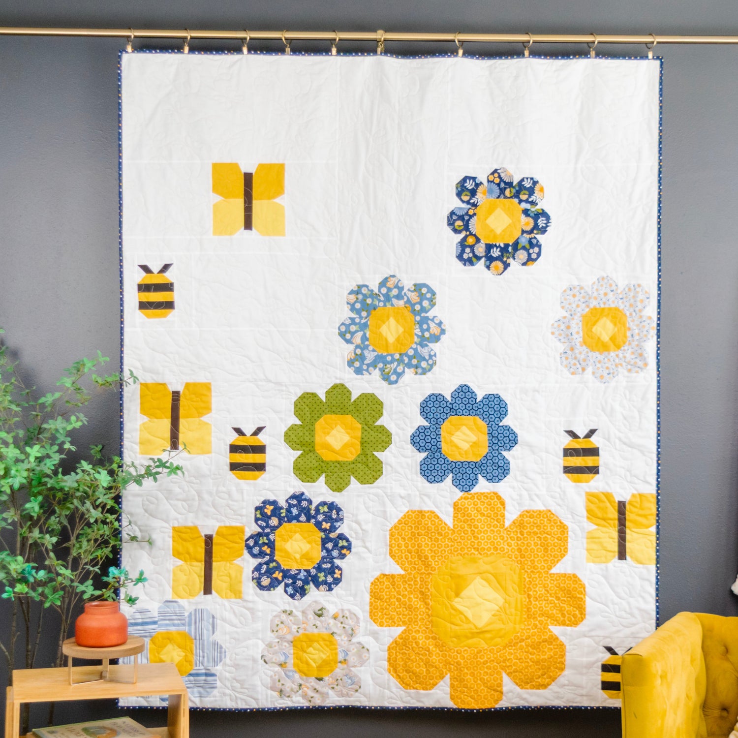 A white quilted wall hanging or blanket featuring colorful floral and bee applique motifs, displayed hanging from a brass rod against a dark gray wall in a styled room with a plant and chair visible.
